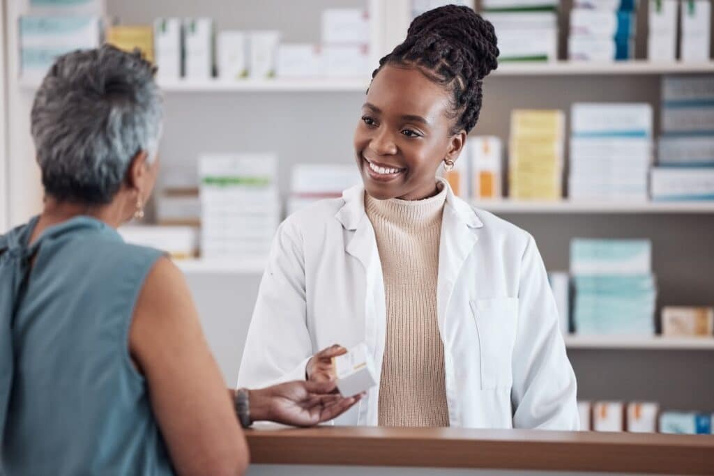 A smiling female pharmacist, dressed in a white coat, assists a customer at the pharmacy counter, handing over a package. The shelves behind her are stocked with various pharmaceutical products.
