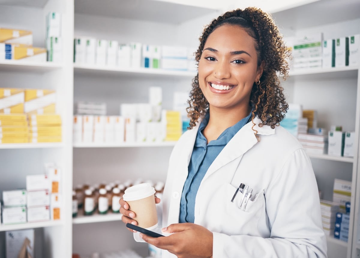 A smiling female pharmacy technician, wearing a white coat and holding a coffee cup and smartphone, stands in a well-stocked pharmacy.
