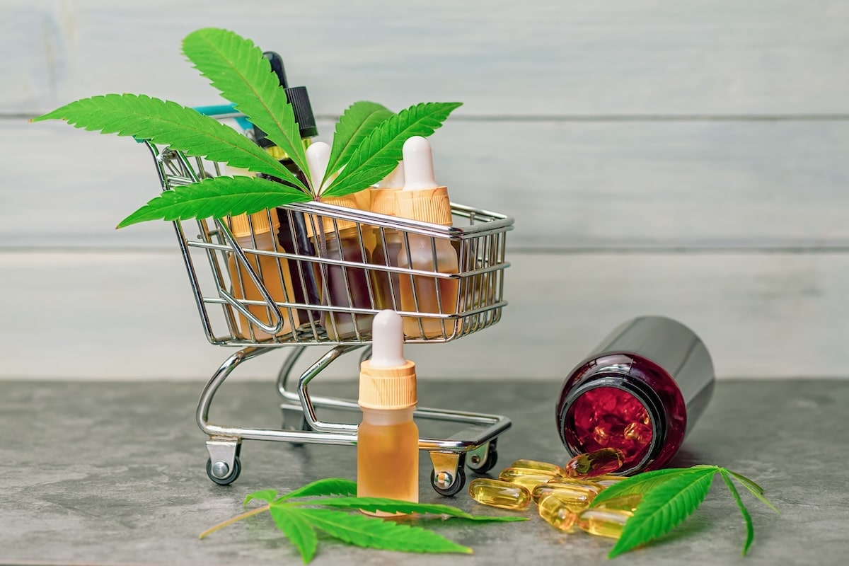 A small shopping cart filled with cannabis CBD oil bottles and cannabis leaves, alongside a spilled bottle of gel capsules on a countertop.
