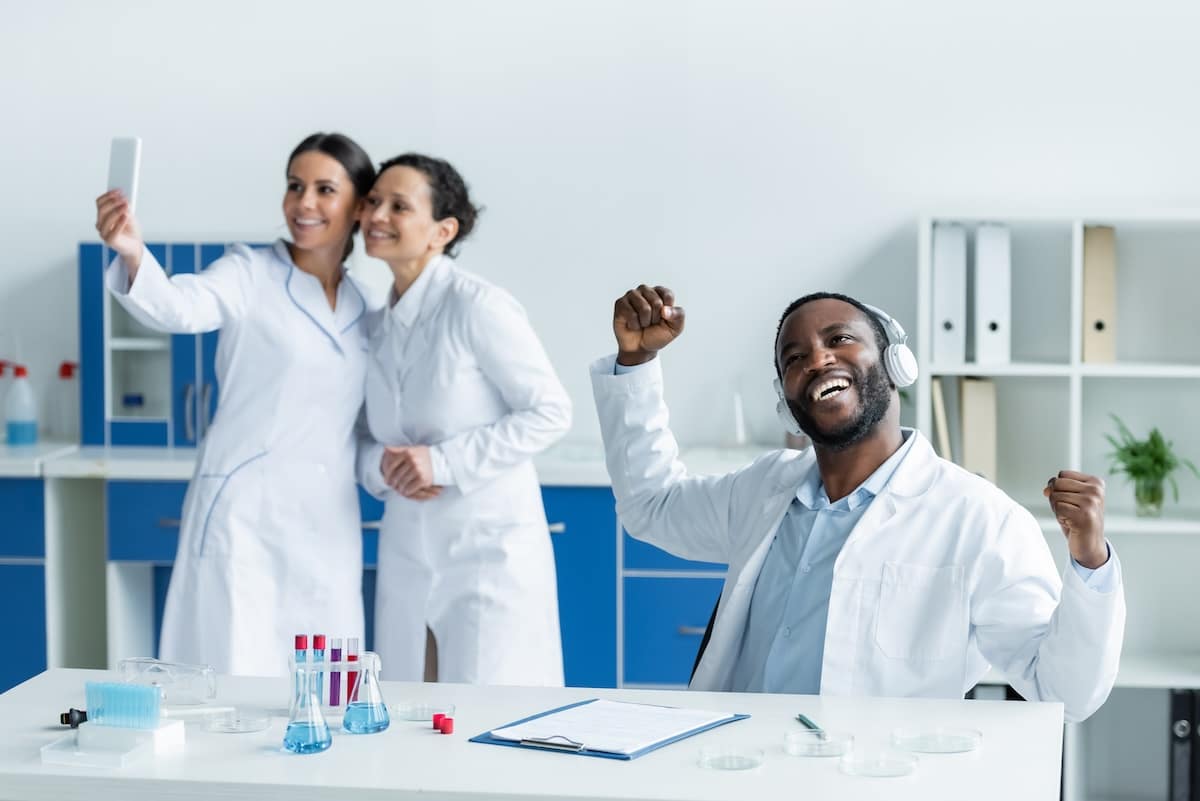 Pharmacist in headphones showing yes gesture near blurred colleagues with smartphone