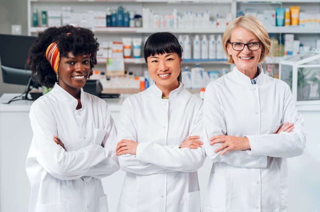 Three successful interracial female pharmacists and medical workers standing in a drugstore with arms crossed and smiling at the camera. A team of professional pharmacists is standing in a pharmacy.