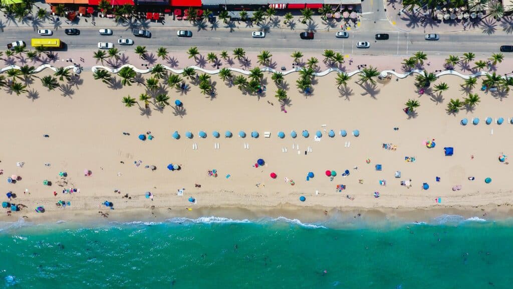 Aerial view of a sandy beach with blue umbrellas, sunbathers, and ocean waves. A row of palm trees and vehicles on a road are visible along the top edge.